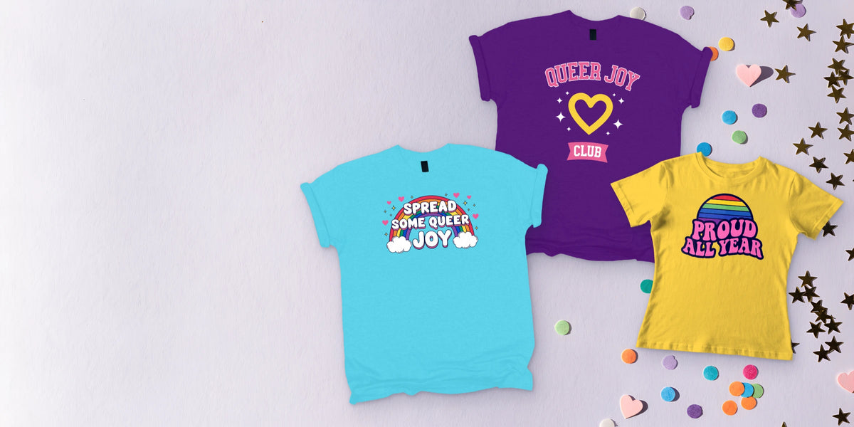 Custom pride T-shirts laid flat on lilac with rainbow confetti: Spread Some Queer Joy, Proud All Year and Queer Joy Club prints