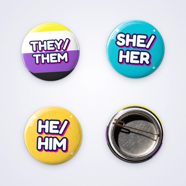 Custom 25mm D-pin pronoun badges reading they/them, she/her and he/him, shown with the D-pin back